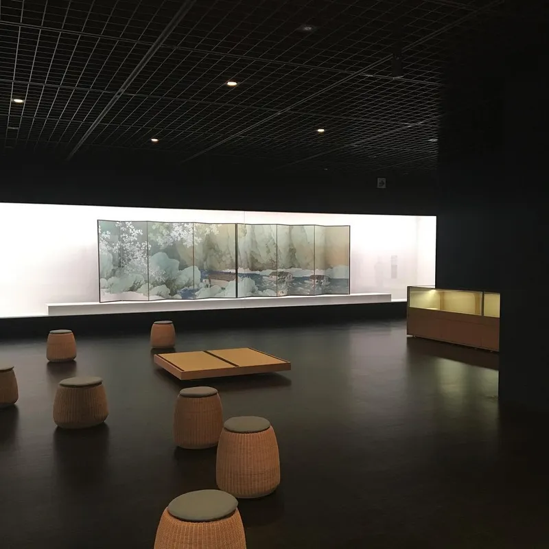 A dimly lit museum gallery with wicker stools facing a backlit Japanese folding screen painting of mountains and cherry blossoms