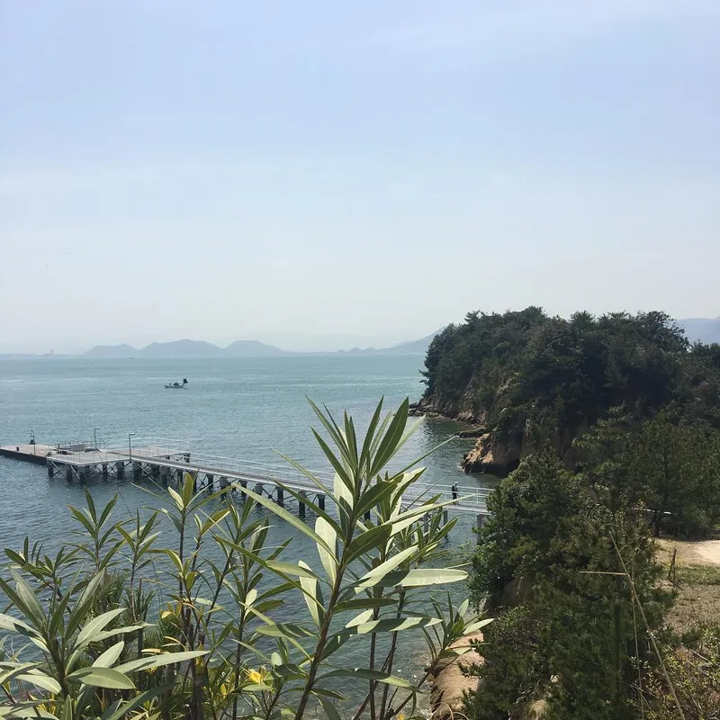 A pier extending into the calm Seto Inland Sea with a wooded rocky island and hazy mountains beyond