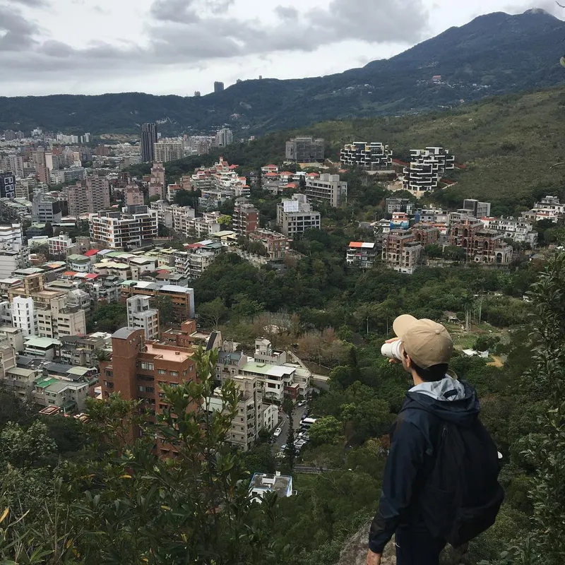 A person in a cap photographing the dense cityscape of Taipei from a hilltop trail
