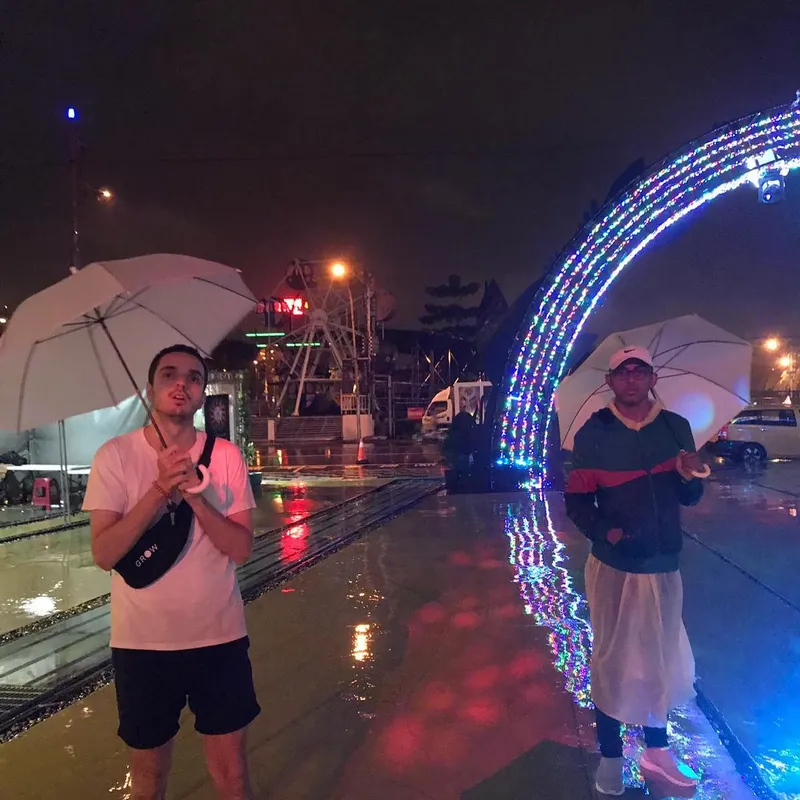 Two men holding white umbrellas in the rain beside a colourful LED arch at night