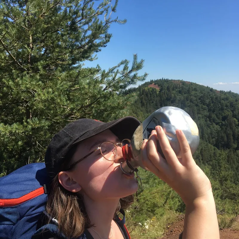 A person in a cap and glasses drinking from a metal flask on a mountain trail