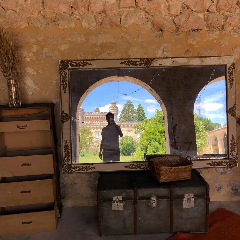 A mirror selfie inside a rustic stone building, reflecting a château and arched windows behind the photographer
