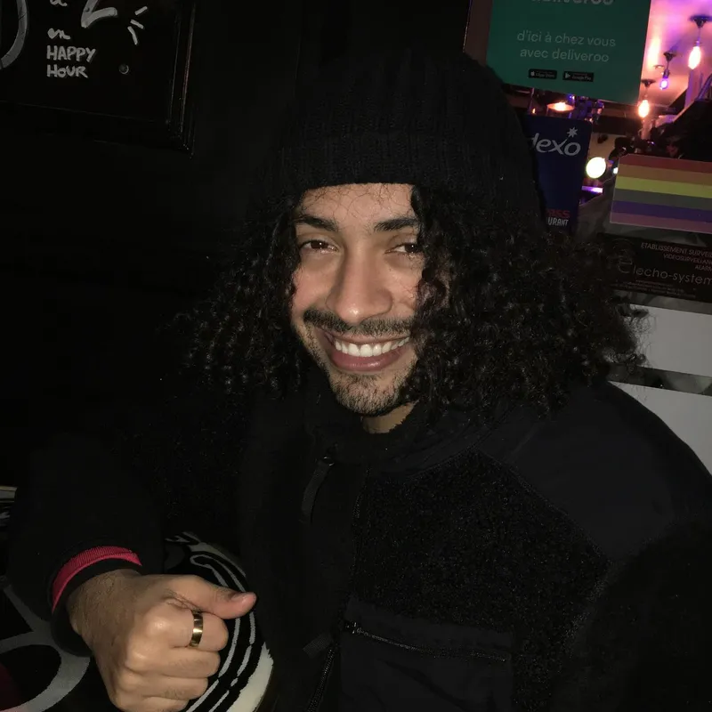 A smiling man with long curly hair and a beanie giving a thumbs up in a bar