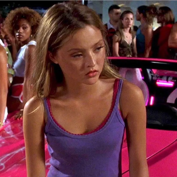 A young woman in a purple tank top standing beside a pink car at a nighttime event