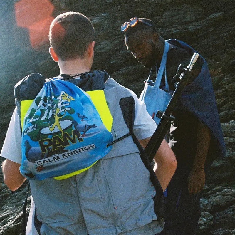 Two people on rocky terrain, one carrying a PAM Calm Energy drawstring bag and the other holding a tripod