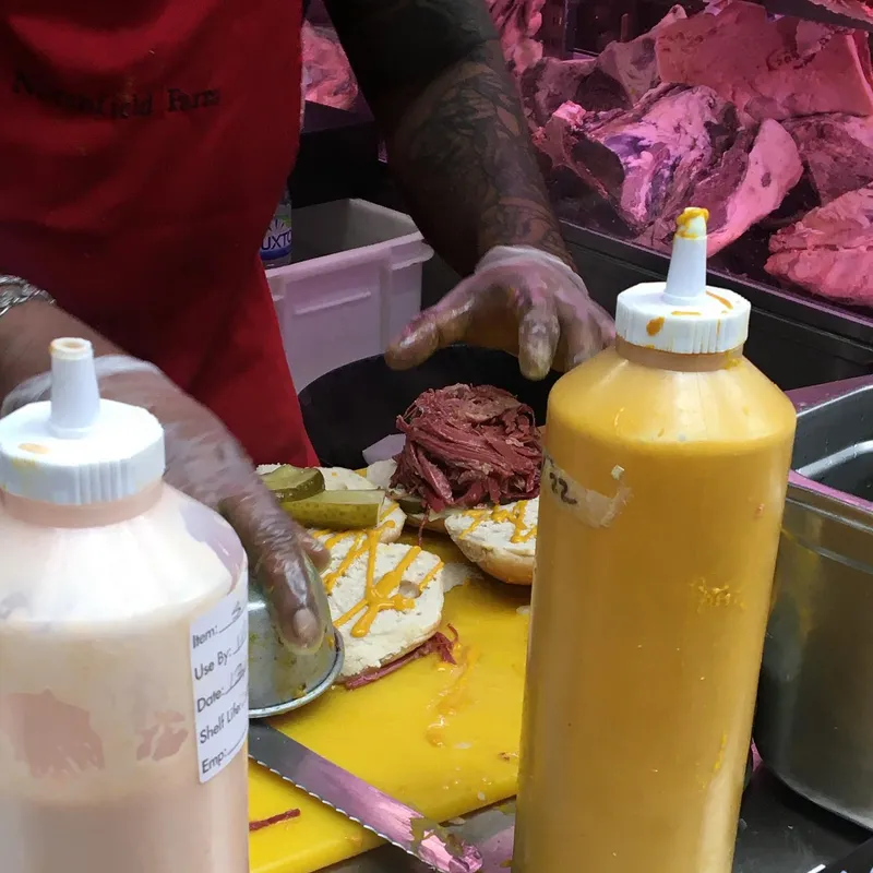 A vendor assembling a salt beef bagel with mustard and pickles at a market stall