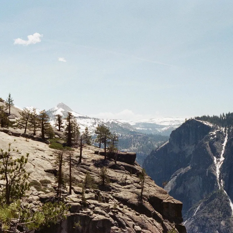 A granite cliff edge with sparse pines overlooking a snowy mountain valley
