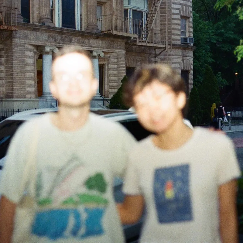 Two out-of-focus friends in graphic tees on a city street in front of a brownstone
