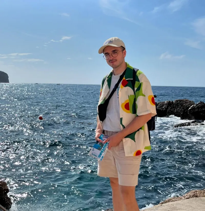 A man in a patterned shirt standing on a rocky coastline with the sea behind him