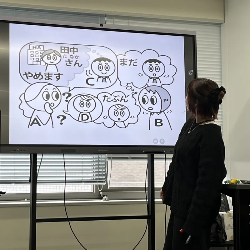 A Japanese language teacher standing before a whiteboard displaying a comic-style dialogue exercise with cartoon characters
