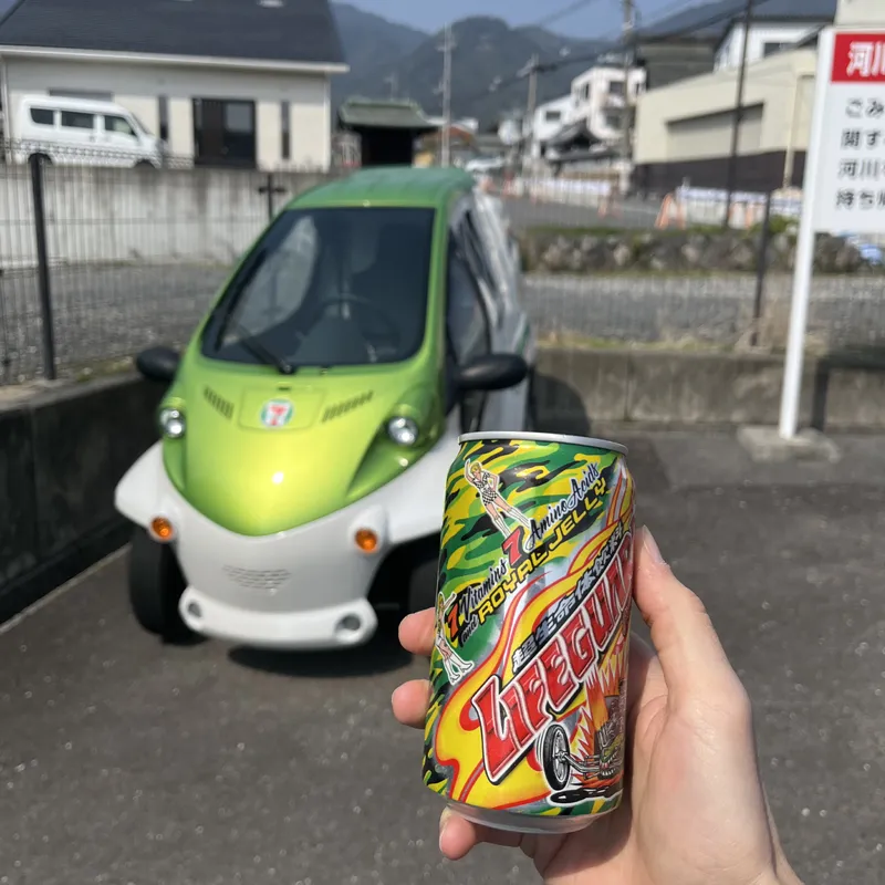 A hand holding a Japanese Lifeguard energy drink can in front of a small lime-green three-wheeled electric microcar with a 7-Eleven logo