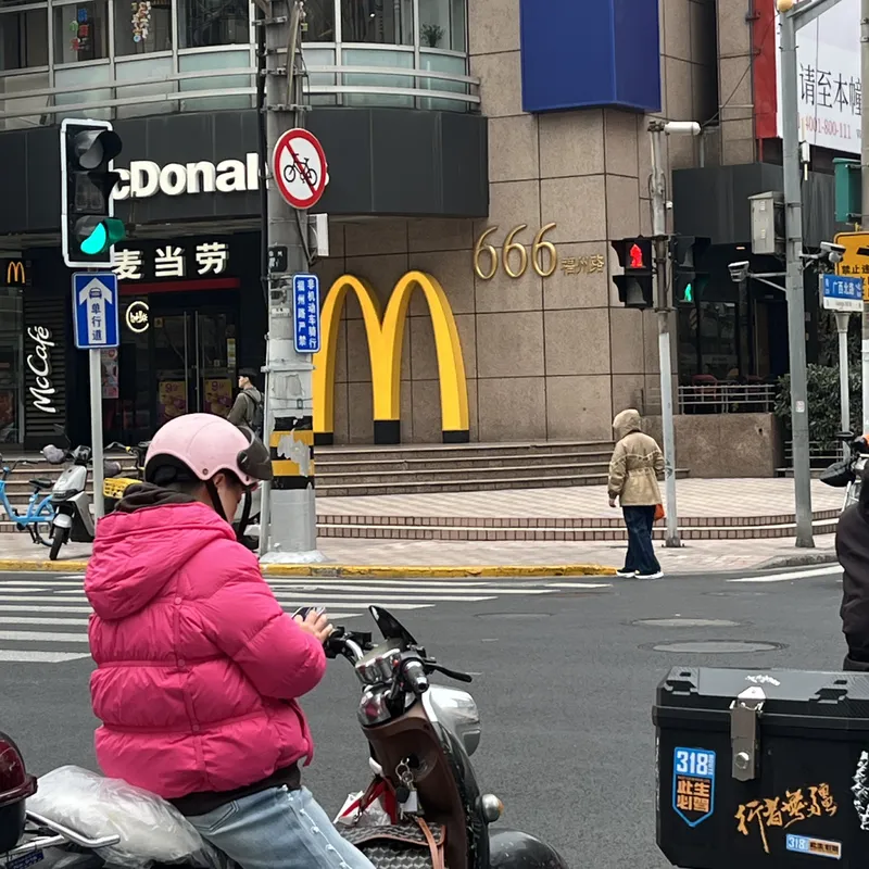 A scooter rider in a pink jacket waiting at a Chinese street intersection with a McDonald's golden arches sign behind her tha the 666 of the street