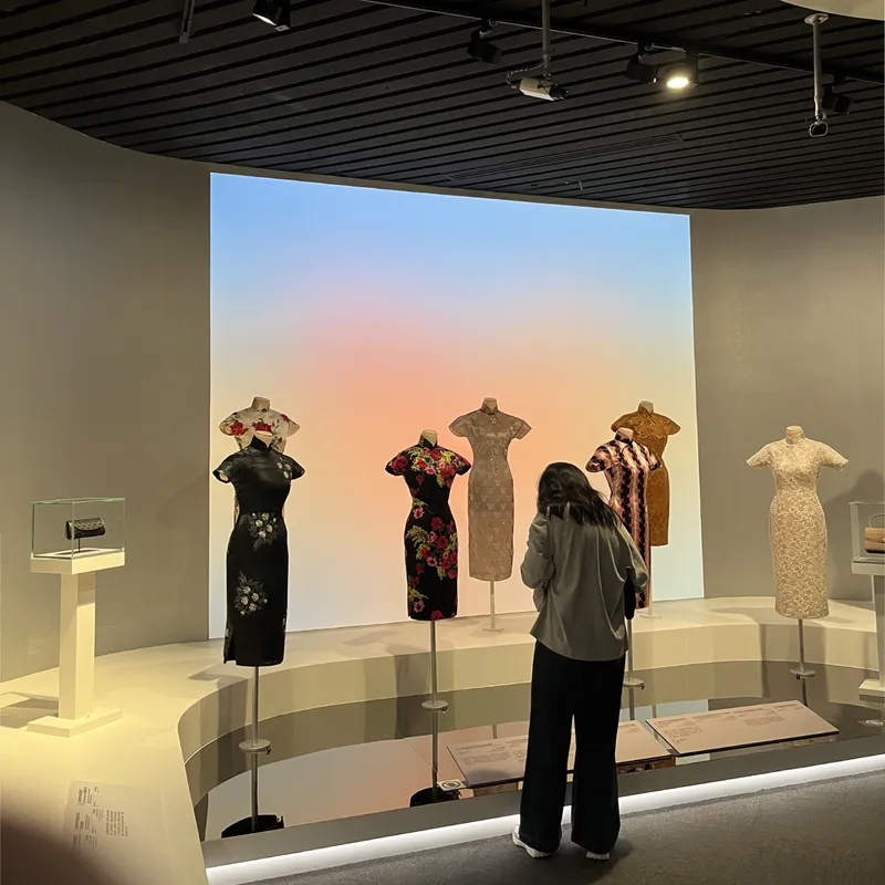 A museum visitor viewing a display of traditional Chinese qipao dresses on mannequins against a soft gradient backdrop