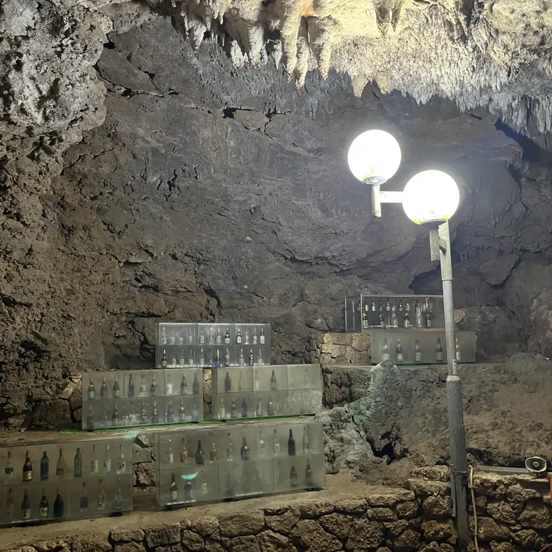 Rows of awamori bottles in glass cases inside a lit natural cave with stalactite ceiling