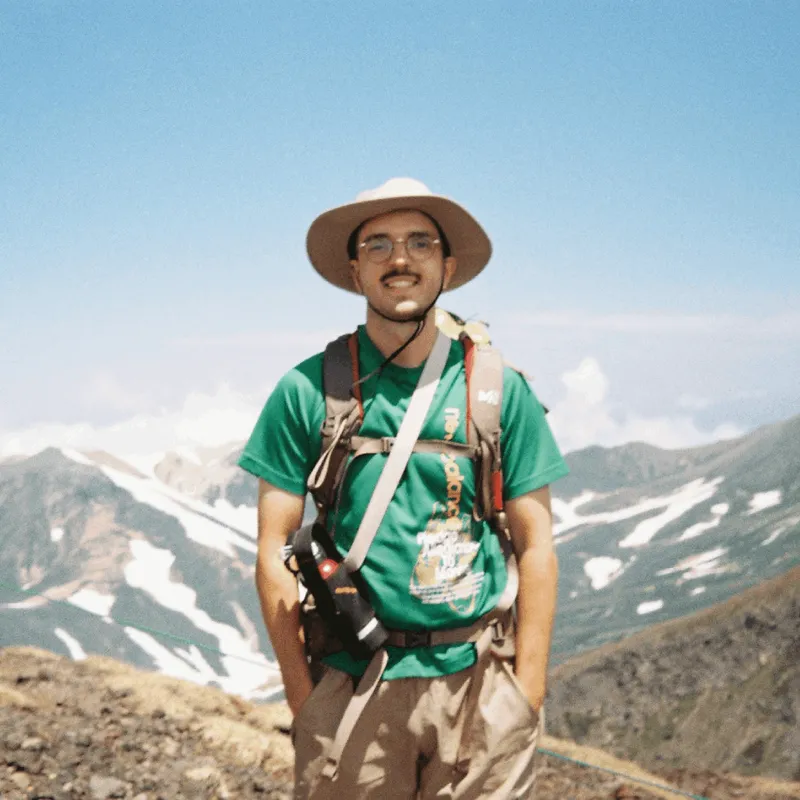 A man wearing a green t-shirt and hiking gear in front of a mountain range